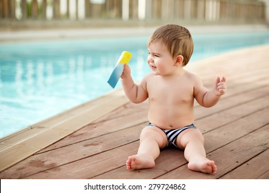 Little Baby Boy Sitting Near The Pool And Is Holding The Sunscreen