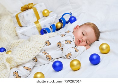 Little Baby Boy Sleeping In Pajamas With A Knitted Plaid On The Background Of Gift Boxes And Colored Balls.