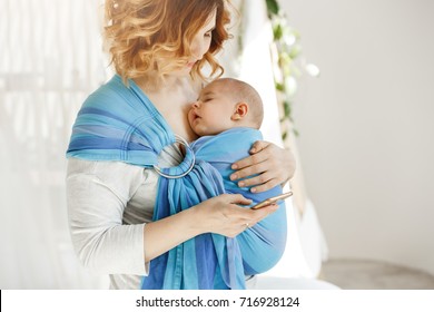 Little Baby Boy Sleeping Tight With Open Mouth And Sweet Face Expression In Baby Sling While Mom Resting Reading World News On Phone. Family Concept.