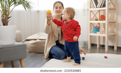 Little baby boy taking colorful toys from his mother and walking on soft carpet in living room. Baby development, family playing games, making first steps, parenthood and care. - Powered by Shutterstock