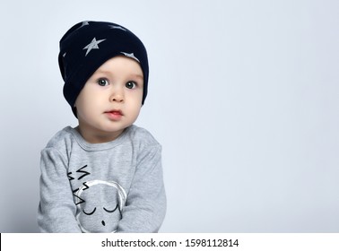 Little Baby Boy Toddler In Grey Casual Jumpsuit, Cap And Barefoot Sitting On Floor And Smiling With Raised Hands Over White Wall Background. Trendy Baby Clothing And Happy Childhood Concept