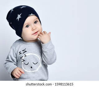 Little Baby Boy Toddler In Grey Casual Jumpsuit, Cap And Barefoot Sitting On Floor And Smiling With Raised Hands Over White Wall Background. Trendy Baby Clothing And Happy Childhood Concept