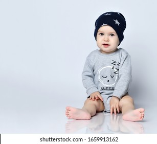 Little Baby Boy Toddler In Grey Casual Jumpsuit, Cap And Barefoot Sitting On Floor And Smiling With Raised Hands Over White Wall Background. Trendy Baby Clothing And Happy Childhood Concept