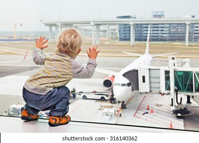 Little Baby Boy Waiting Boarding To Flight In Airport Transit Hall And Looking Through The Window At Airplane Near Departure Gate. Active Family Lifestyle, Travel By Air With Child On Summer Vacation