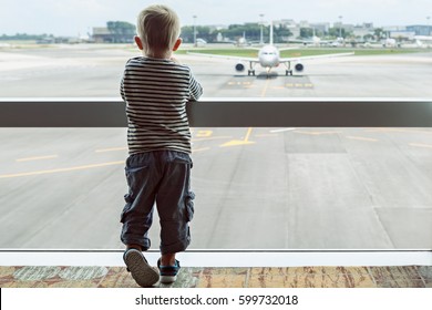 Little Baby Boy Waiting Boarding To Flight In Airport Transit Hall And Looking Through The Window At Airplane Near Departure Gate. Active Family Lifestyle, Travel By Air With Child On Summer Vacation