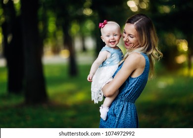 Little Baby Girl And Her Mother Walking In The Park At The Day Time. Baby Is Looking At The Camera And Smiling, Showing Two Funny White Teeth. Mother Holding Her And Smiling Too.