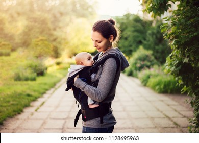 Little baby girl and her mother walking outside during sunset. Mother is holding and tickling her baby, babywearing in the ergo carrier - Shutterstock ID 1128695963