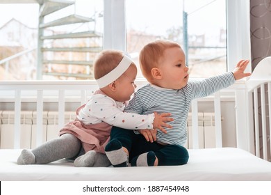 A little baby girl hugs a baby boy while sitting together in a white crib. There is a window in the background. The Concept Of Valentine's Day.