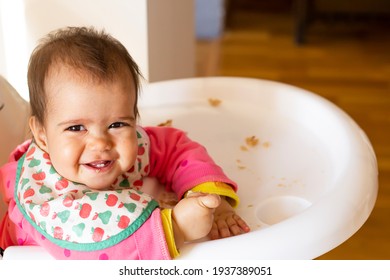 Little Baby Girl Is Smiling And Eating Piece Of Bread. Baby Eating Sitting On Highchair At Home. Baby Girl 10-12 Months Is Eating A Wheat Bread At Breakfast Table. The Baby Collects Crumbs Of Bread.