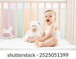 little baby girl smiling or laughing in the crib at home in the nursery in a white bodysuit, cute funny baby playing with soft and wooden toys before going to bed, lifestyle