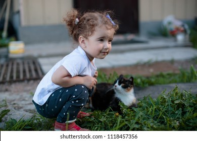 Little Baby Girl Squatting Next To A Cat In The Yard