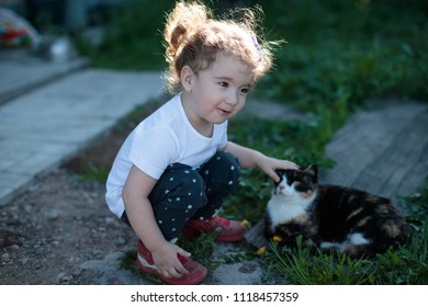 Little Baby Girl Squatting Next To A Cat In The Yard