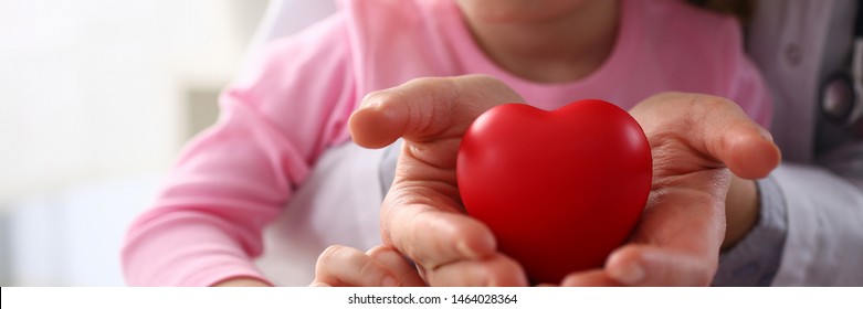 Little Baby Girl Visiting Doctor Holding In Hands Red Toy Heart As Life Safe Symbol