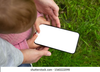 Little Baby Kid Toddler Holding Smart Phone With Hands Of Adult Parent. Blank Screen. Kid Play Gave, Soft The Net, Learning By The Mobile Phone In Hands Of A Child. Glass Outside On The Background. 