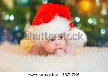 Little baby in red cap of Santa Claus celebrates Christmas. Christmas photo of infant in red cap. New Year's holidays and Christmas tree.