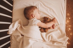 A Little Baby In A White Bodysuit Sleeping In A White Crib Holding A Teddy Bear And Surrounded By Small Lights