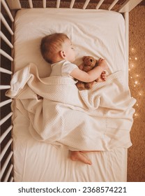 A little baby in a white bodysuit sleeping in a white crib holding a teddy bear and surrounded by small lights