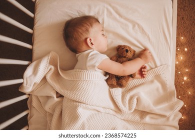 A little baby in a white bodysuit sleeping in a white crib holding a teddy bear and surrounded by small lights - Powered by Shutterstock