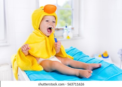 Little Baby In Yellow Duck Towel Brushing Teeth On Changing Table After Bath. Infant Boy With Tooth Brush. Dental Hygiene, Toothbrush And Toothpaste For Young Kids. Child Teeth And Oral Health Care