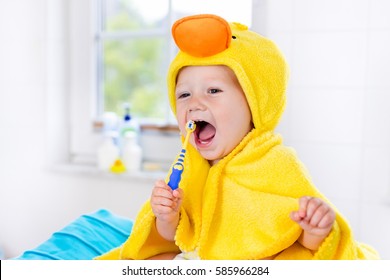 Little Baby In Yellow Duck Towel Brushing Teeth On Changing Table After Bath. Infant Boy With Tooth Brush. Dental Hygiene, Toothbrush And Toothpaste For Young Kids. Child Teeth And Oral Health Care.