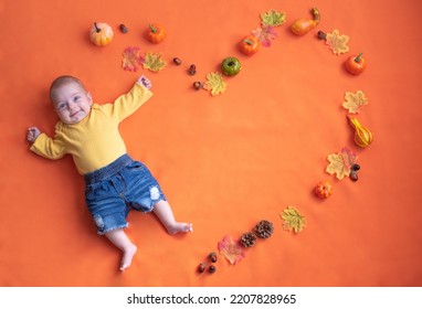 Little Baby In Yellow Suit And Jeans. Newborn Baby On Orange Background Decorate Pumpkin, Autumn Leaves Shape Of Heart. Top View. Holiday Halloween, Love Family, Beautiful Card. Copy Space