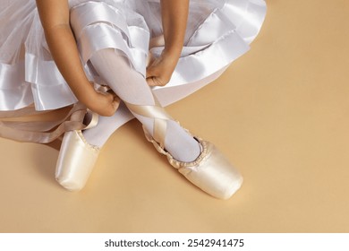 Little ballerina tying pointe shoes on beige background, closeup - Powered by Shutterstock