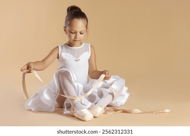 Little ballerina tying pointe shoes on beige background, space for text - Powered by Shutterstock