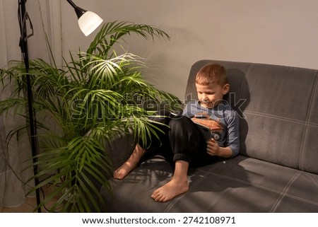Little barefoot boy using tablet alone on grey sofa at night with floor lamp and tropical houseplant in dark cozy room. Concept of child screen addiction, digital dependency and late night device use.