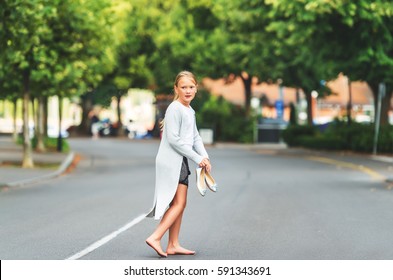 Little barefoot girl walking down the street, wearing long grey cardigan, holding silver bow ballerinas shoes