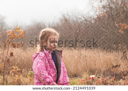 Little beautiful cute sad girl in a jacket with blond hair in pigtails stands on an autumn background