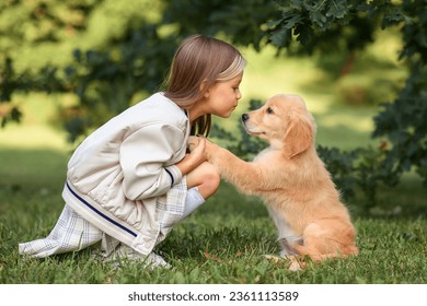 a little beautiful girl in a beige jacket and a plaid skirt with small golden retriever puppies dogs plays in the park in summer - Powered by Shutterstock