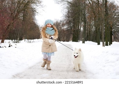 Little Beautiful Girl, Cute Happy Positive Child Kid Is Walking At Snow, Snowy Day Having Fun With Her Dog On A Leash At Winter Day In Park Or Forest