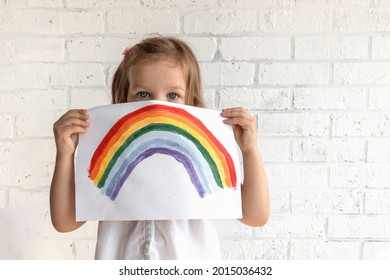 A little beautiful girl looks at the camera   holds sheet paper which rainbow is drawn  Pandemic hope concept  Stay at home