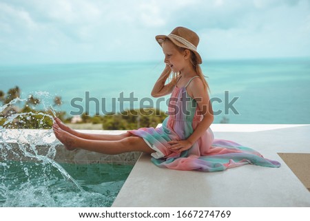 Little and beautiful girl with red hair and freckles sits by the pool in the villa and poses in front of the camera. Children's fashion