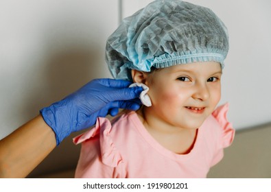 Little Beautiful Girl Smiles And Looks Into The Camera. Waiting For The Doctor To Clean The Ear And Make A Puncture. Child Piercing.