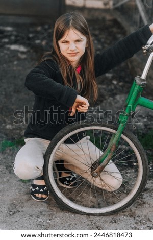 Little beautiful sad teenage girl, dissatisfied upset child showing dislike thumbs down sits near an old retro bicycle with a broken, punctured wheel tire outdoors. Photography, portrait, lifestyle.