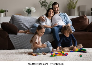 Little biracial kids have fun sit on floor play with toys building bricks together. Smiling multiracial parents relax on couch hugging enjoying. Happy multiethnic family with children rest at home. - Powered by Shutterstock