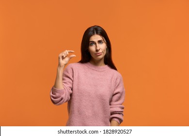 A little bit. Beautiful woman shows little bit gesture, asks some more, please give me few more. shows that this is insignificant. wears in pink knitted sweater isolated over orange background