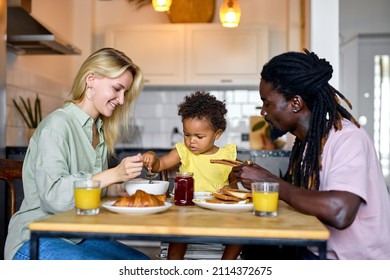 Little Black Baby Sitting In High Chair And Eating Food With Parents Indoors. Baby's First Feeding. Cute Toddler Holding Spoon. In The Morning, In Cozy Kitchen. Diverse Man And Woman With Child