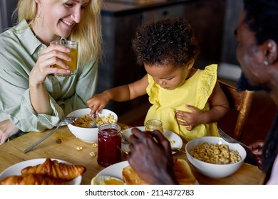 Little Black Baby Sitting In High Chair And Eating Food With Parents Indoors. Baby's First Feeding. Cute Toddler Holding Spoon. In The Morning, In Cozy Kitchen. Diverse Man And Woman With Child