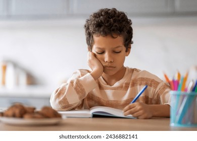 Little black boy feeling tired and sleepy while doing his homework, exhausted african american preteen schoolboy sitting at desk in kitchen and falling asleep while writing in notebook, copy space