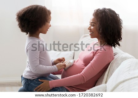 Little Black Girl Placing Baby Shoes At Pregnant Mom's Belly While They Sitting Together On Couch In Living Room, Playing And Bonding, Side View