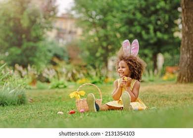 Little Black girl wear bunny ears and gathering Easter eggs on Easter egg hunt in garden - Shutterstock ID 2131354103