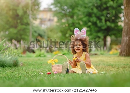 Little Black girl wear bunny ears and gathering Easter eggs on Easter egg hunt in garden