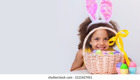 Little Black girl in yellow dress with pink bunny ears paint Easter eggs and hiding behind Basket full of colored eggs