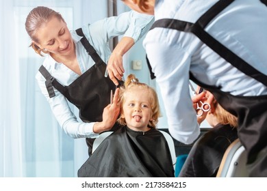 Little blond 3 years old boy stick out tongue while hairdresser woman make a haircut looking at the mirror