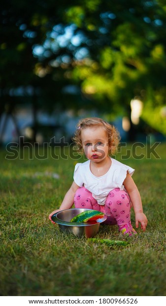 Little blond baby with surprised face\
in crouching pose with pan of watermelon in\
park