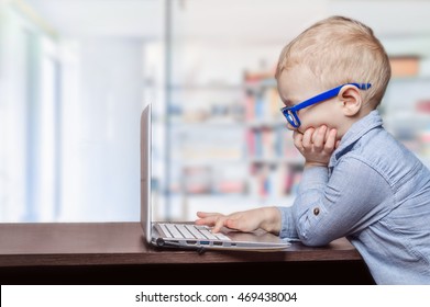  little blond boy in the blue glasses and blue shirt sitting at a computer Desk looking at the laptop screen.  concept of modern education