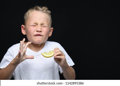 Little, blond boy is eating a piece of a lemon in front of black background and making a facial expression.