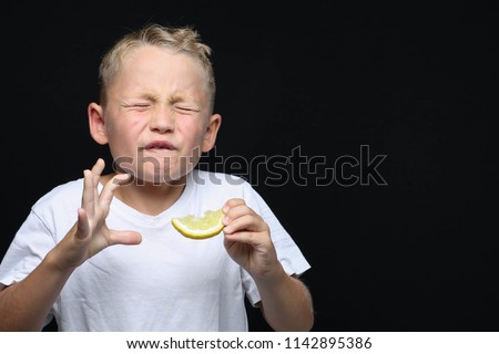 Little, blond boy is eating a piece of a lemon in front of black background and making a facial expression.